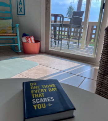 A book on a table titled Do Something Everyday that Scares You with a coastal furnishings and a view of a balcony with sunlight filtering in.