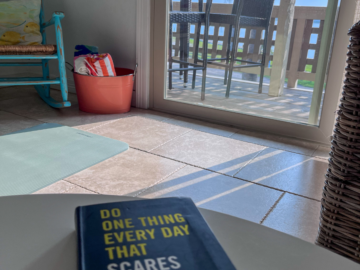 A book on a table titled Do Something Everyday that Scares You with a coastal furnishings and a view of a balcony with sunlight filtering in.