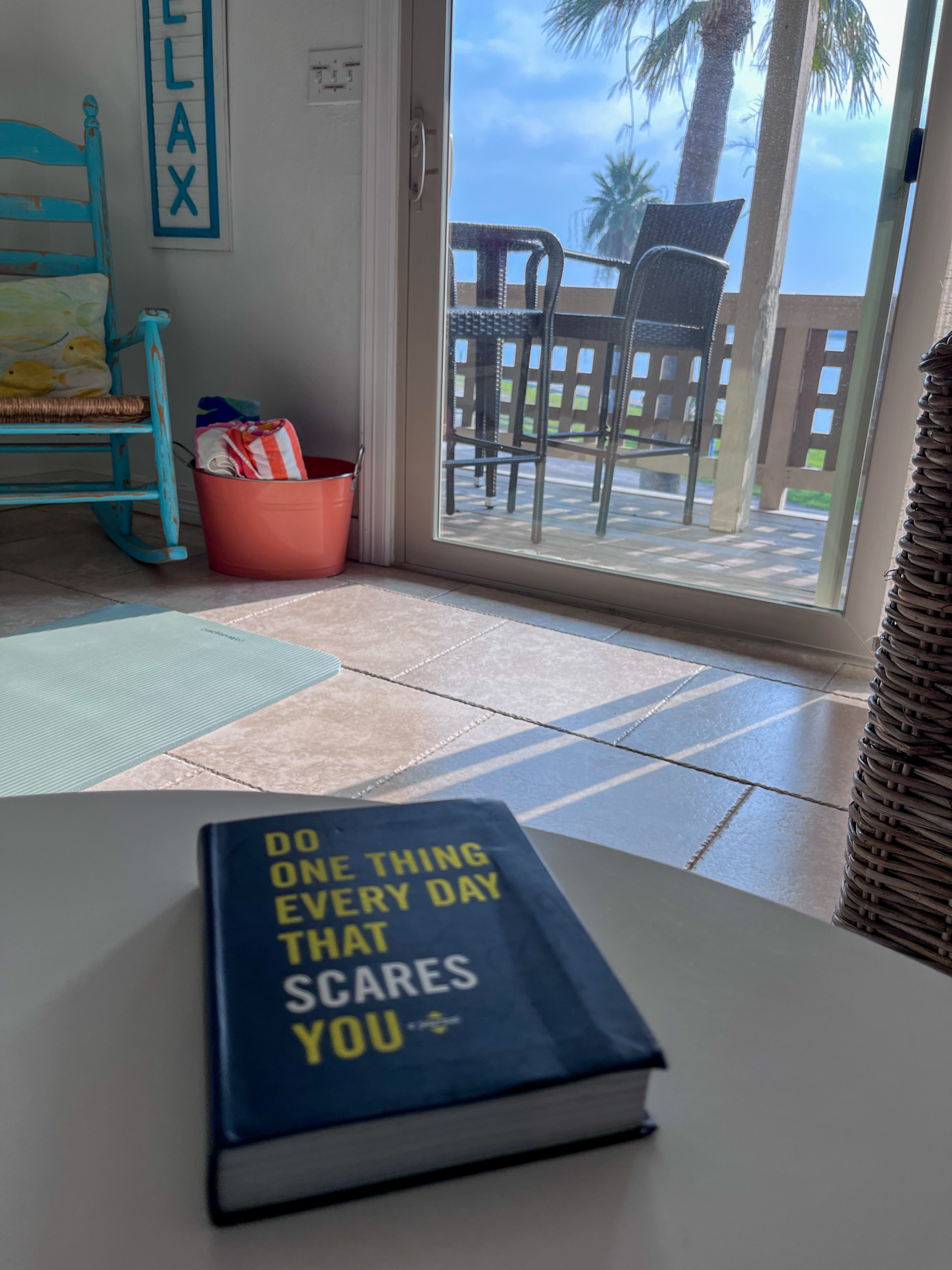 A book on a table titled Do Something Everyday that Scares You with a coastal furnishings and a view of a balcony with sunlight filtering in.