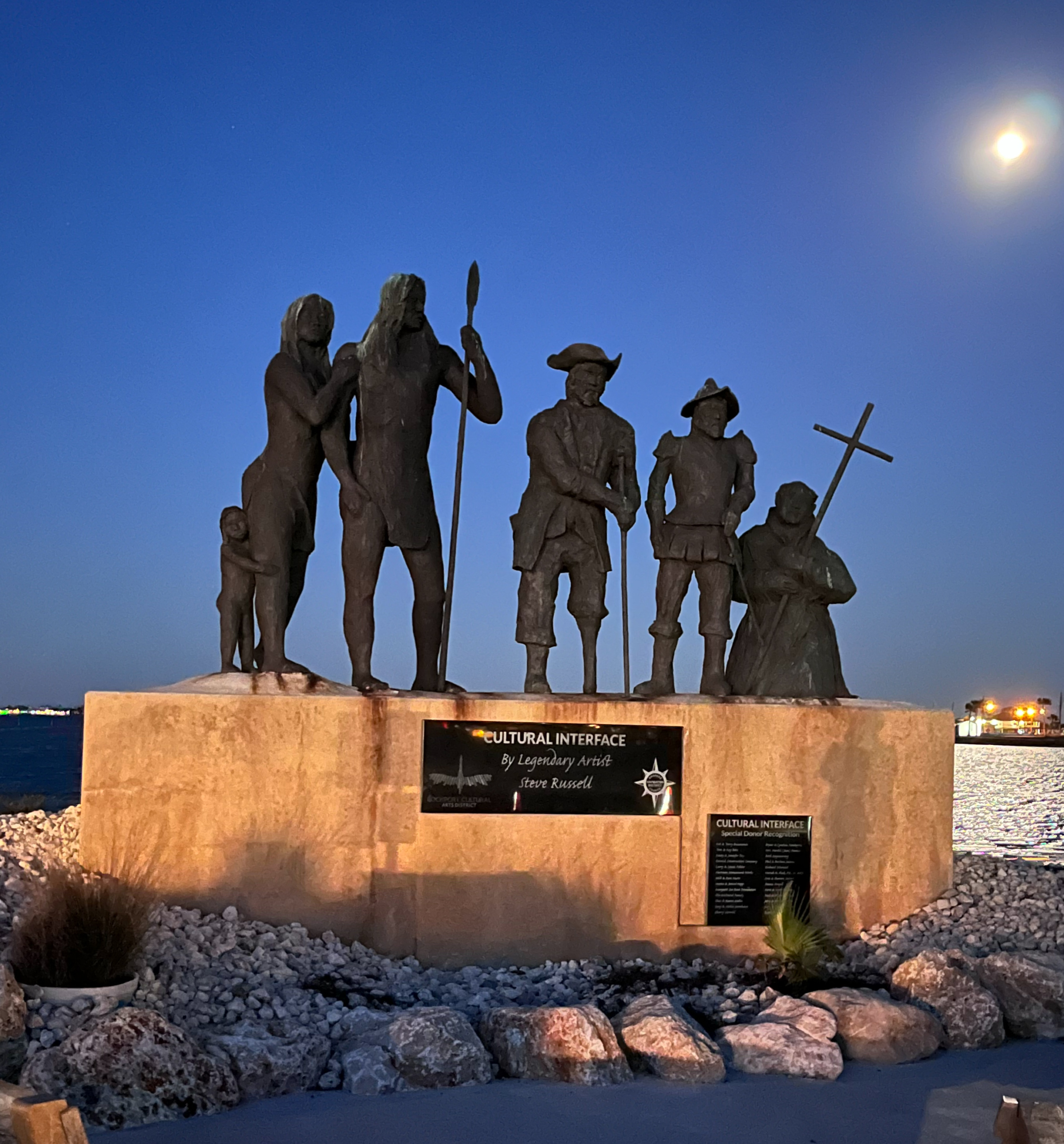 Bronze statue depicting early settlers together on a waterfront monument at night, illuminated under a moonlit sky.